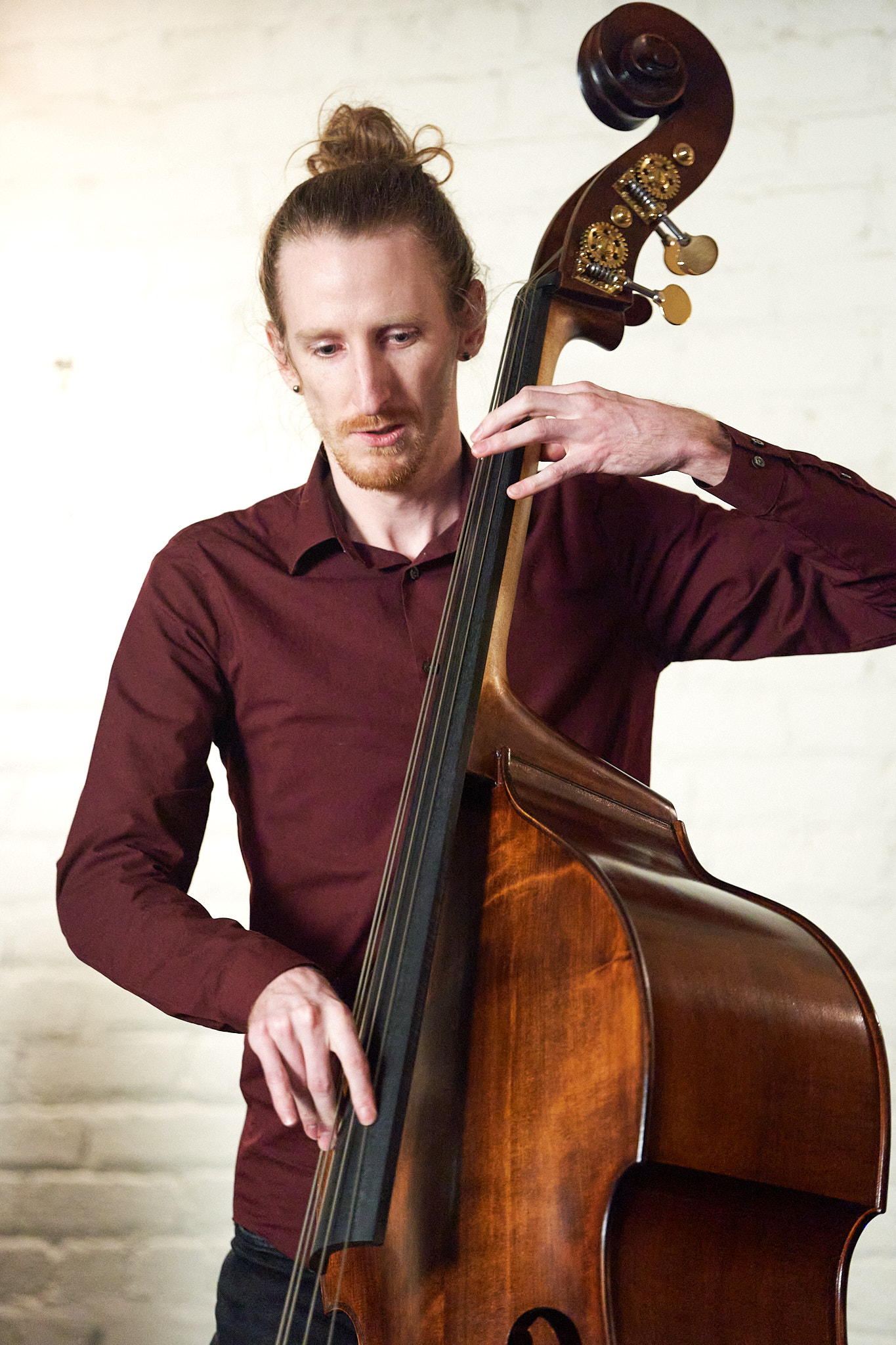 Photo of Sam playing upright bass, close-up, indoors, a white brick wall behind him.